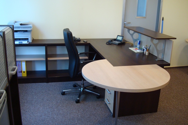 Zdjęcie przedstawia ciemnobrązowe biurko narożne z jasnym, okrągłym blatem pomocniczym, czarne krzesło biurowe na kółkach, regał z półkami oraz drukarkę, całość w biurowym wnętrzu z szarą wykładziną.